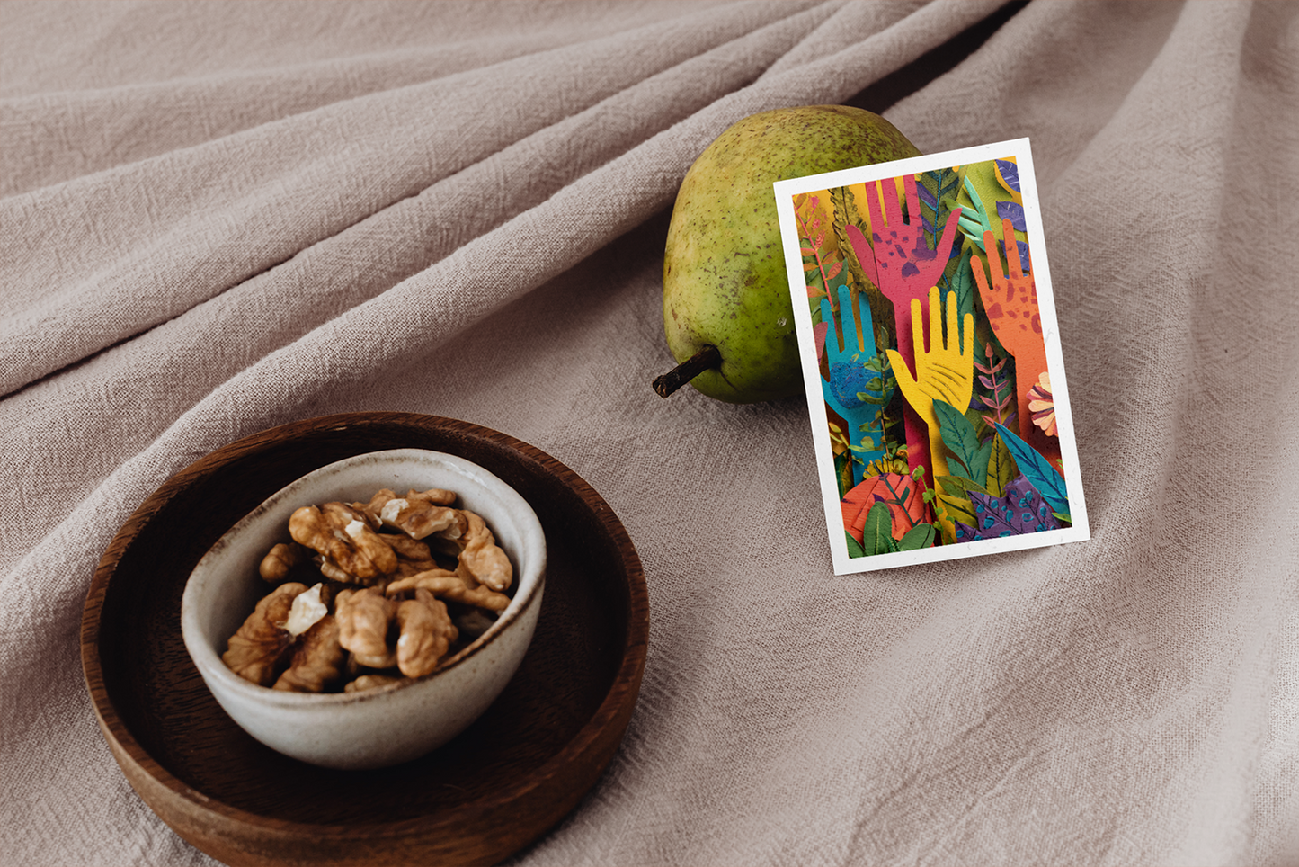 Bowl of nuts, pear, and colorful card with hand designs on a beige fabric background