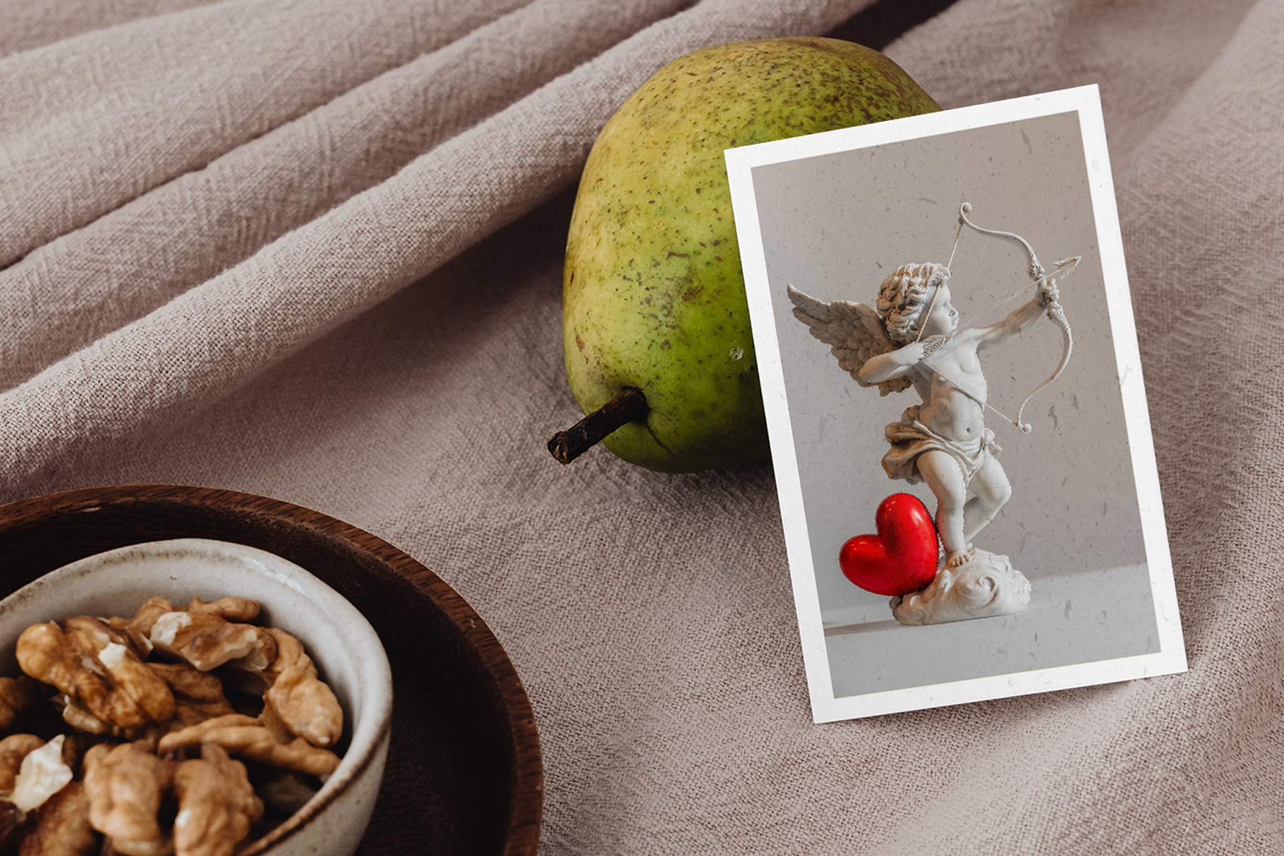 Pear, bowl of nuts, and photo of Cupid with a heart on a beige fabric background