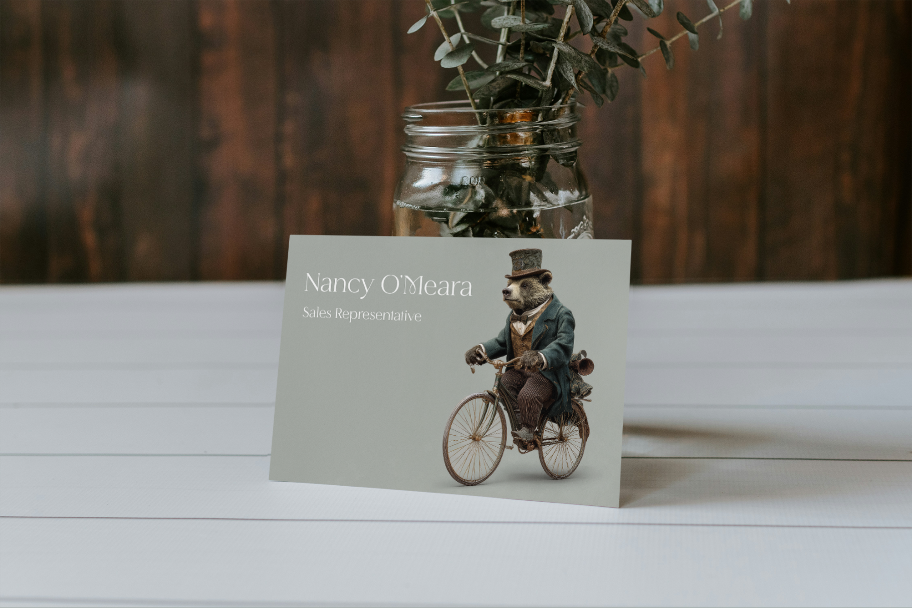 Business card with a bear on a bicycle design on a wooden surface with a vase of plants in the background.