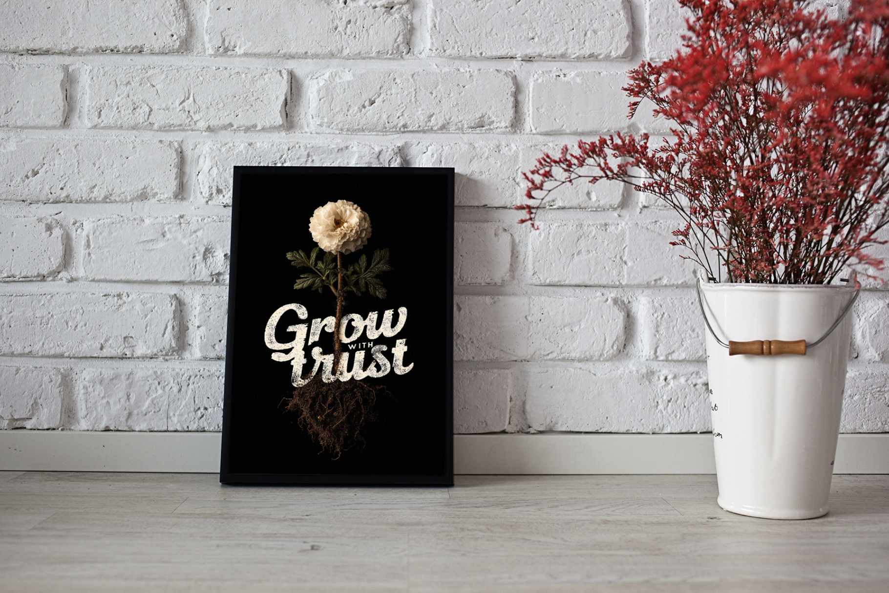 Decorative sign with 'Grow trust' text next to a white bucket with red flowers against a white brick wall.