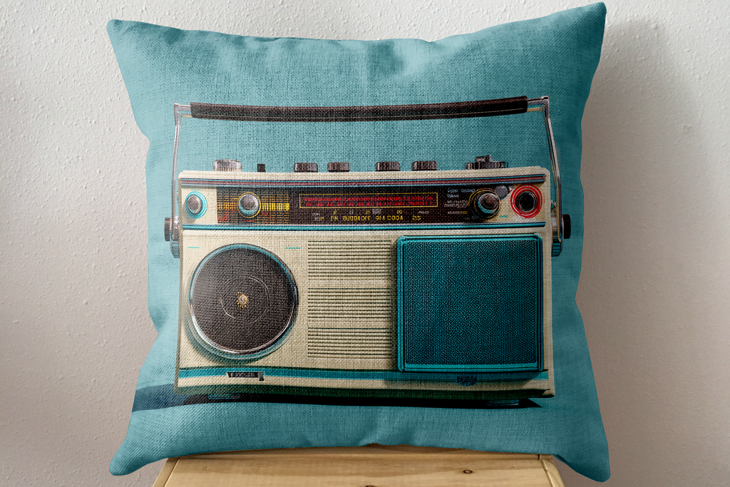 A square teal throw pillow featuring a vintage-style retro radio printed on the front, resting on a light wood surface against a textured off-white wall.