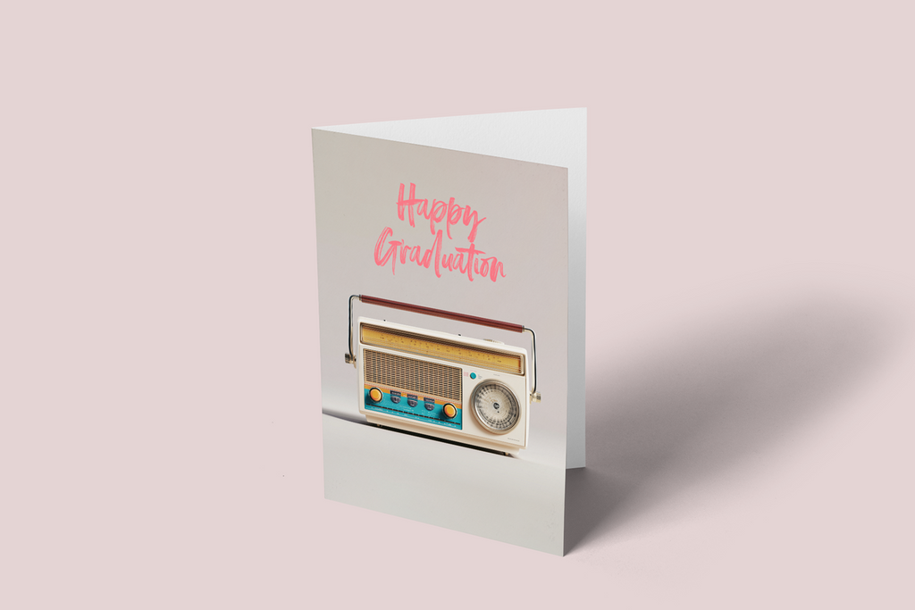 A folded greeting card standing upright on a pale pink background. The card is white with pink script reading 'Happy Graduation' and features a retro radio illustration in cream, blue, yellow, and gray.