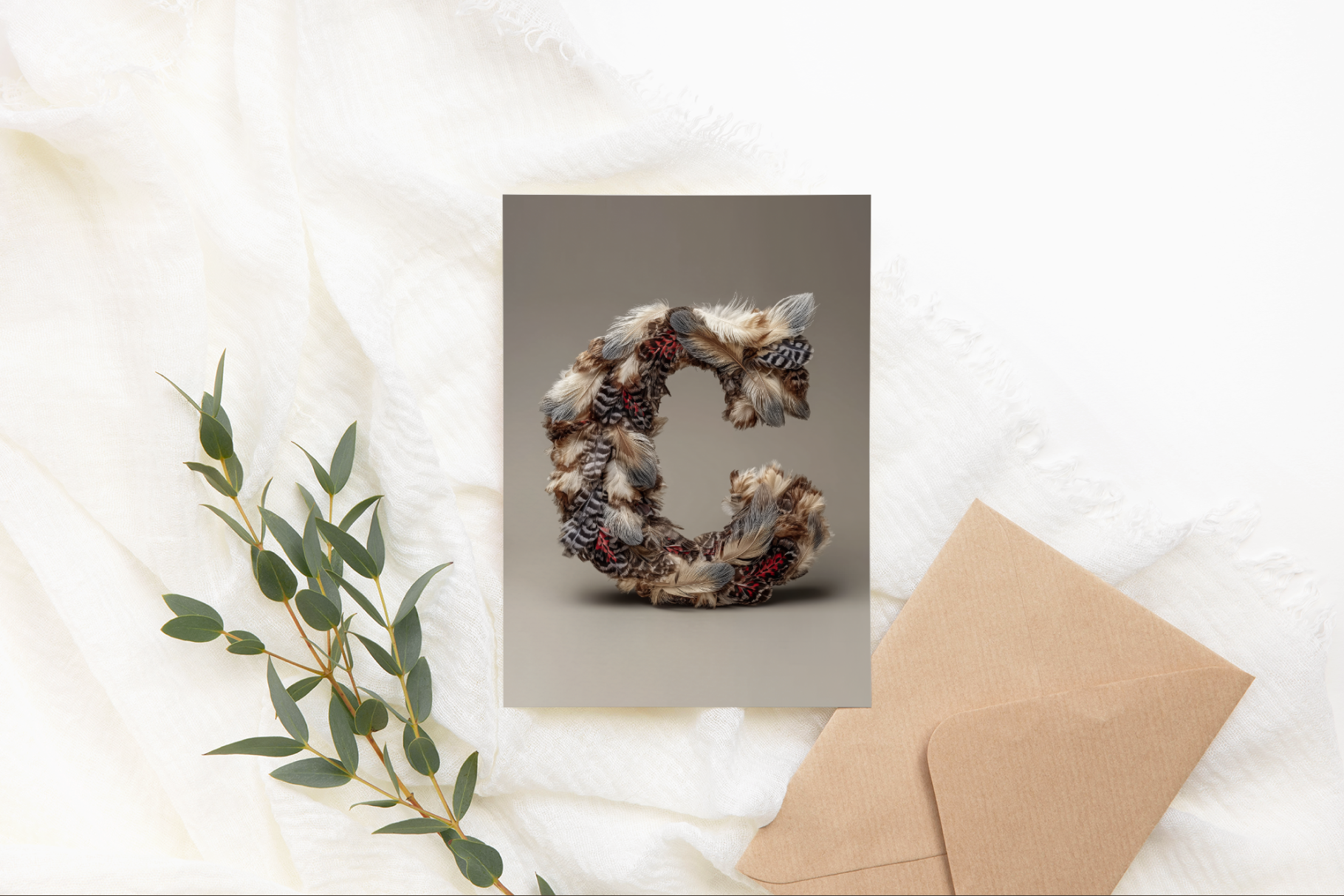 Greeting card with decorative letter 'C' on a textured surface with a brown envelope and greenery.